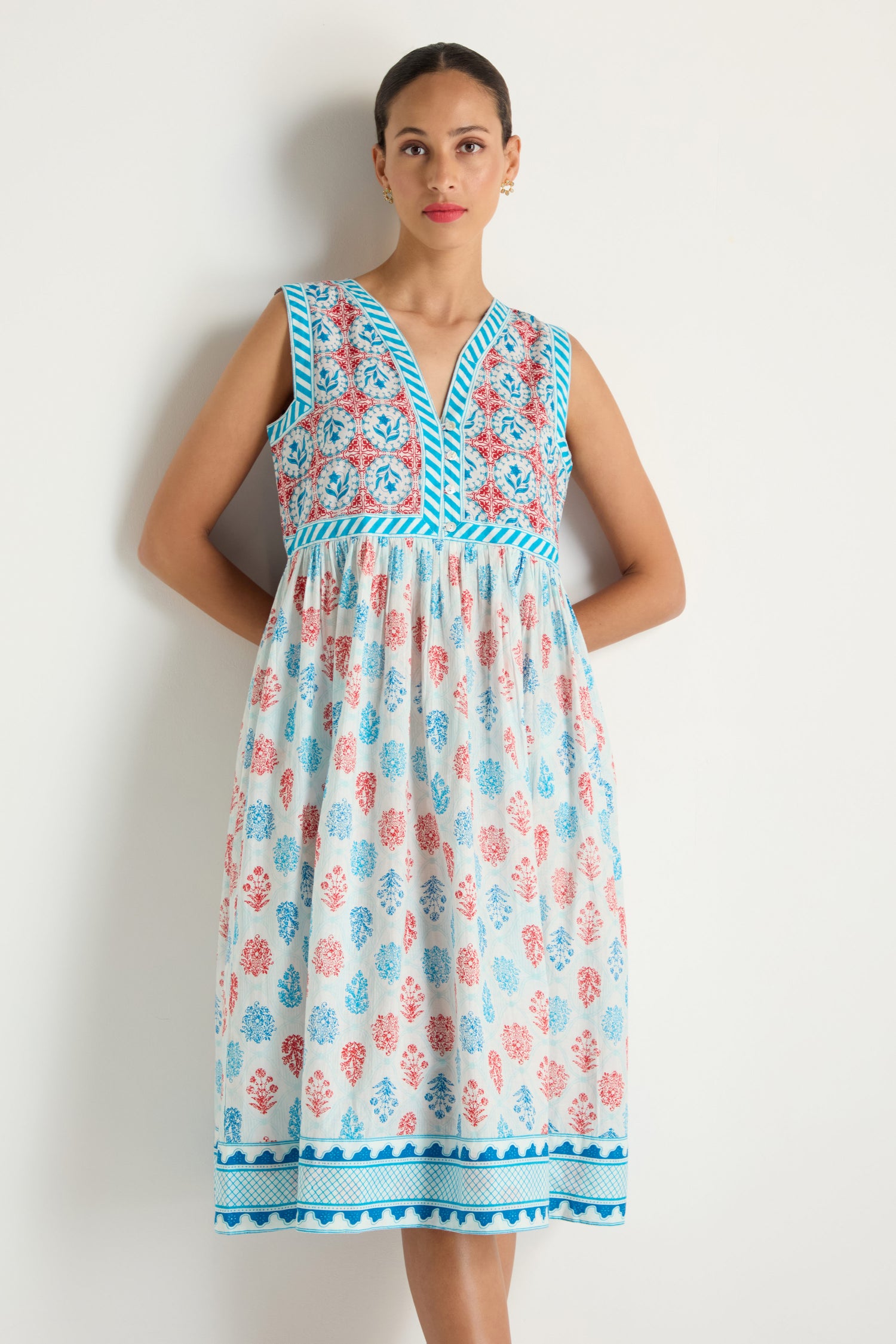 A woman stands against a white wall wearing the Chevron Flared Dress, which is sleeveless and features blue trim, a patterned bodice, and a skirt with red, blue, and pink floral cotton block prints.