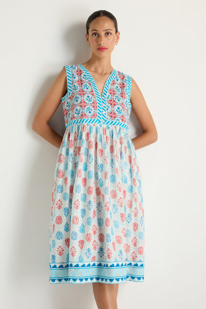 A woman stands against a white wall wearing the Chevron Flared Dress, which is sleeveless and features blue trim, a patterned bodice, and a skirt with red, blue, and pink floral cotton block prints.