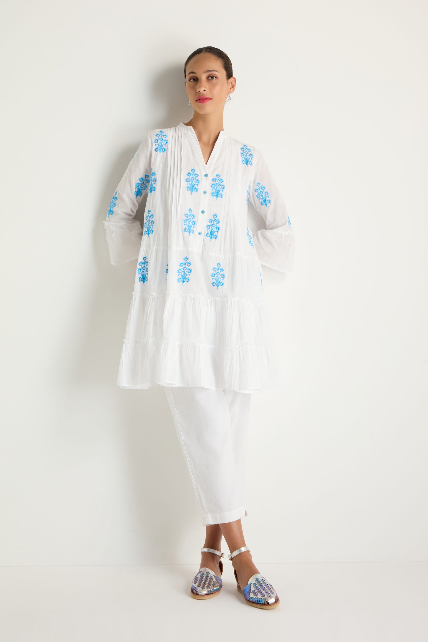 A woman stands against a white wall wearing the Embroidered Floral Flared Sleeve Tunic, styled with white pants and flat sandals.