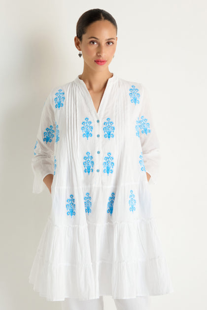 A woman stands against a white background wearing the Embroidered Floral Flared Sleeve Tunic, featuring pure cotton fabric and delicate blue floral designs.