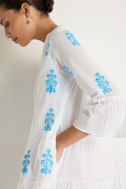 A person in profile wears the Embroidered Floral Flared Sleeve Tunic, a pure cotton white dress with blue floral embroidery, flared sleeves, and textured flowing fabric, set against a plain light background.