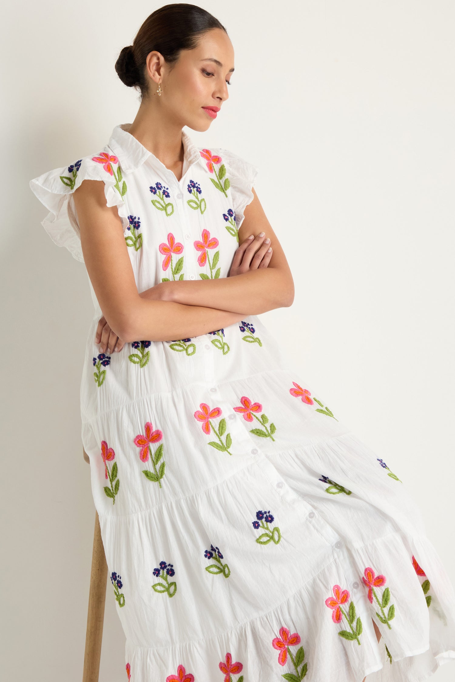 A woman sits on a wooden stool against a light background, wearing the Embroidered Flower Frill Sleeve Dress—a cotton midi dress with colorful floral embroidery and flutter sleeves.