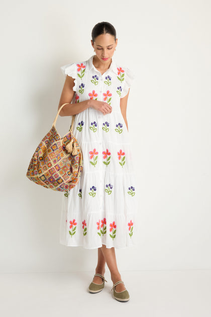 A woman stands against a white wall wearing the Embroidered Flower Frill Sleeve Dress, featuring colorful floral details, and holds a large patterned shoulder bag with beige flat shoes.