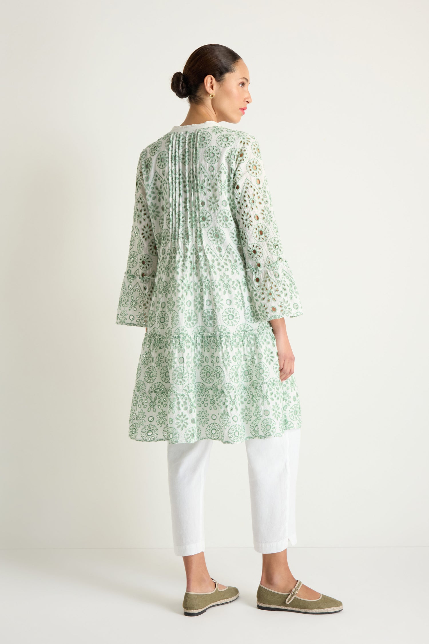 A woman faces away, wearing the Embroidered Circle Flared Sleeve Tunic in light green and white with broderie anglaise embroidery, paired with white pants and green slip-on shoes against a plain background.