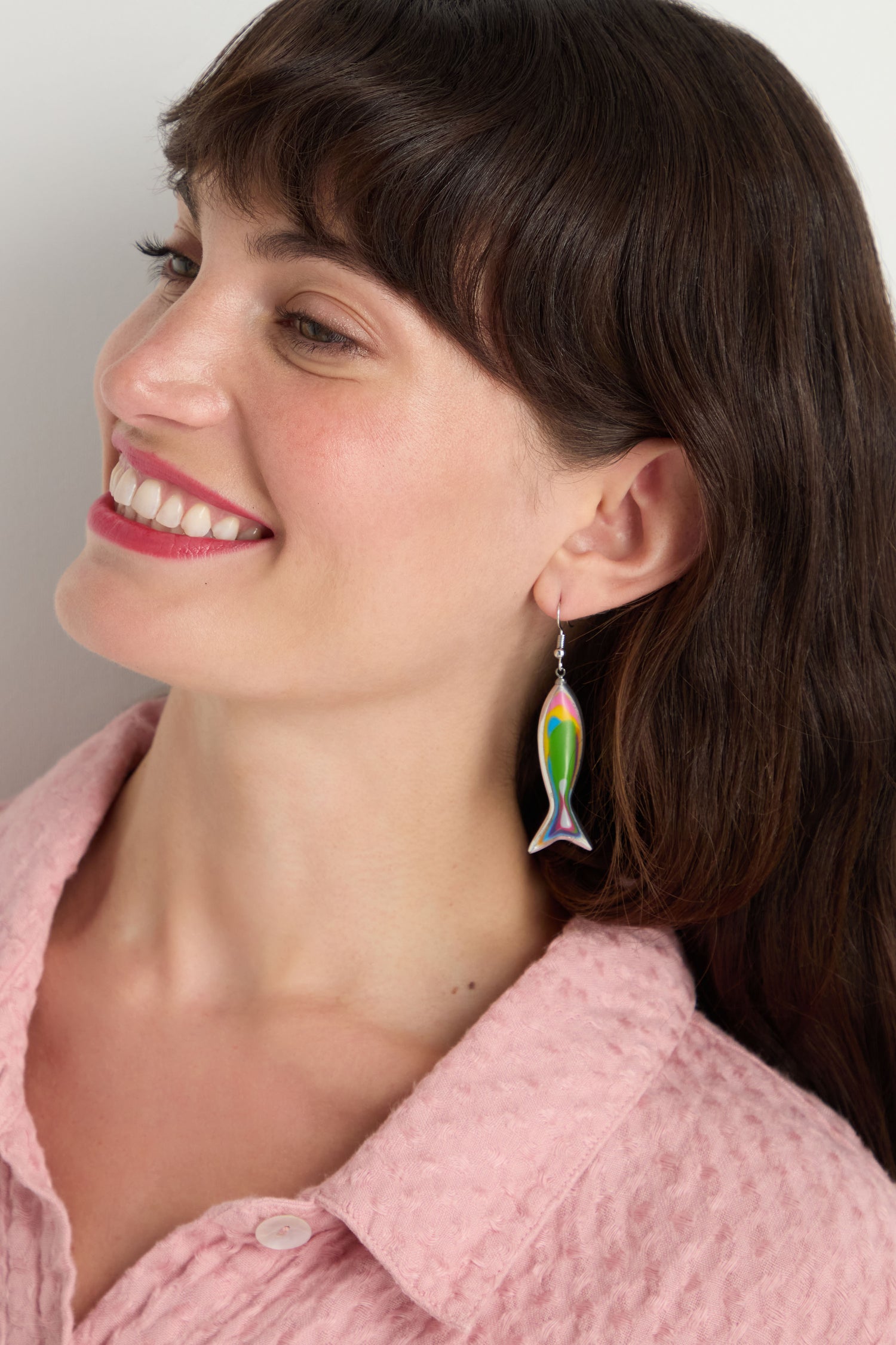 Smiling woman with long brown hair wears a pink textured shirt and vibrant Poisson Earrings, looking to the side.