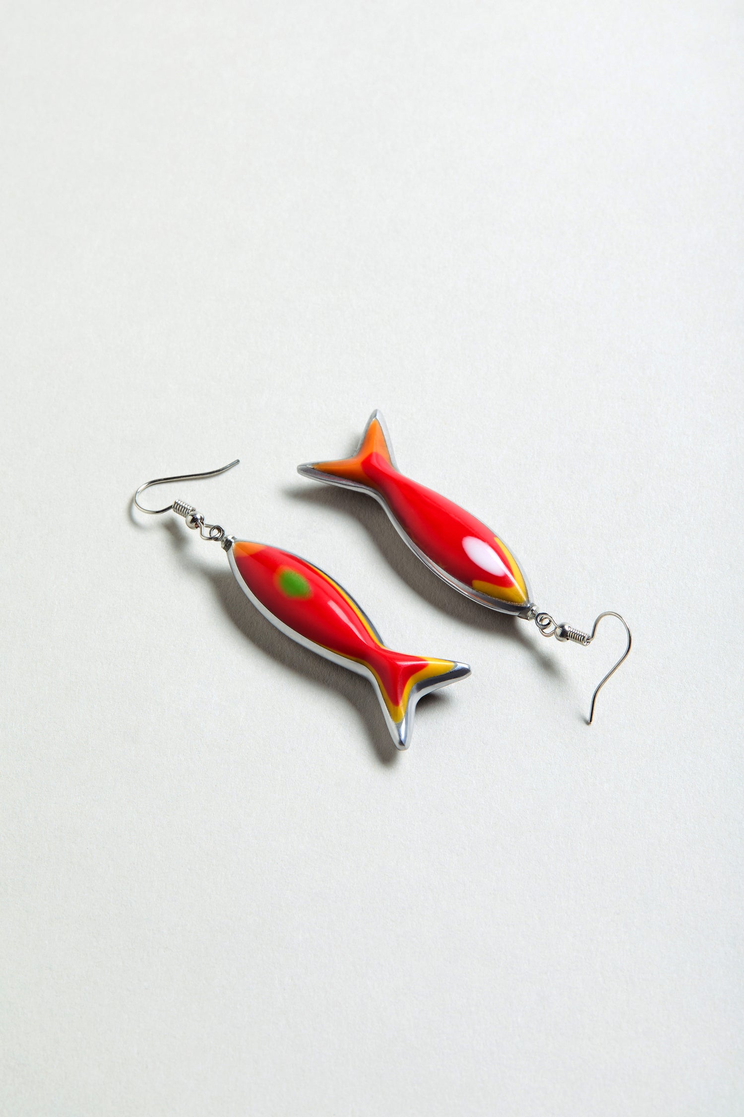 The Poisson Earrings are fish-shaped and crafted from recycled aluminium, featuring red, yellow, and green accents. Displayed on a plain light background.