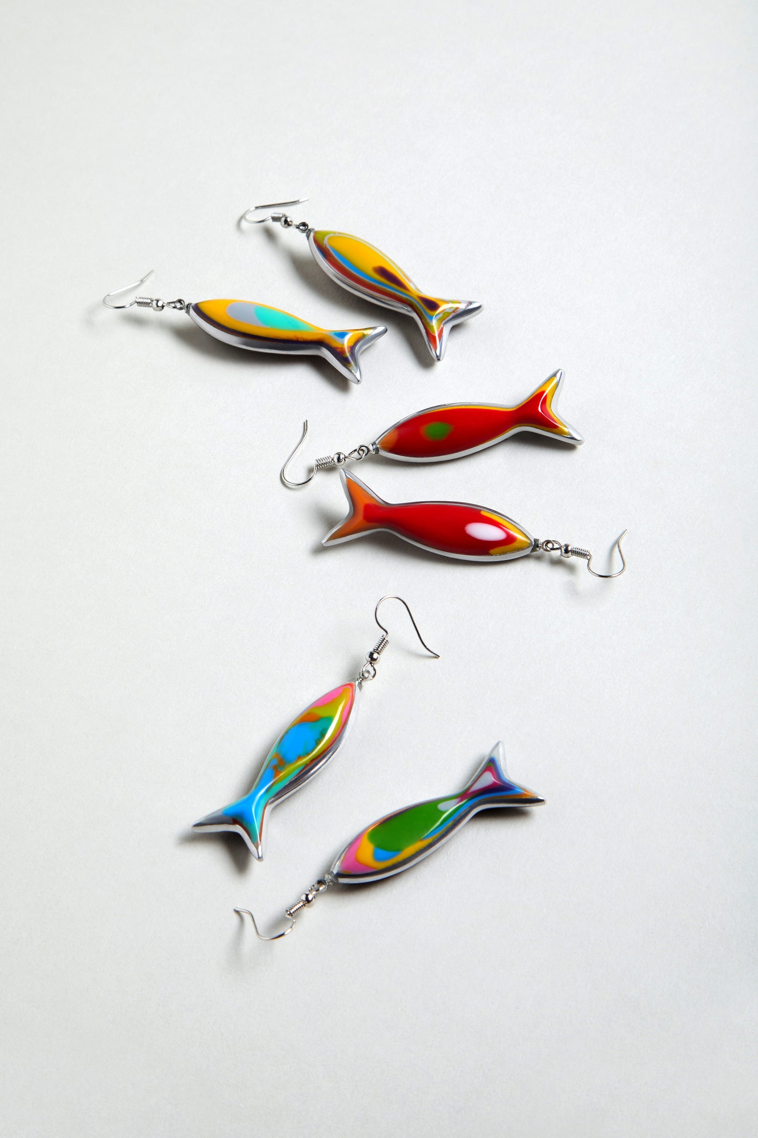 Six vibrant Poisson Earrings, crafted from multicolour resin in playful fish shapes with metal hooks, are displayed on a light grey surface.