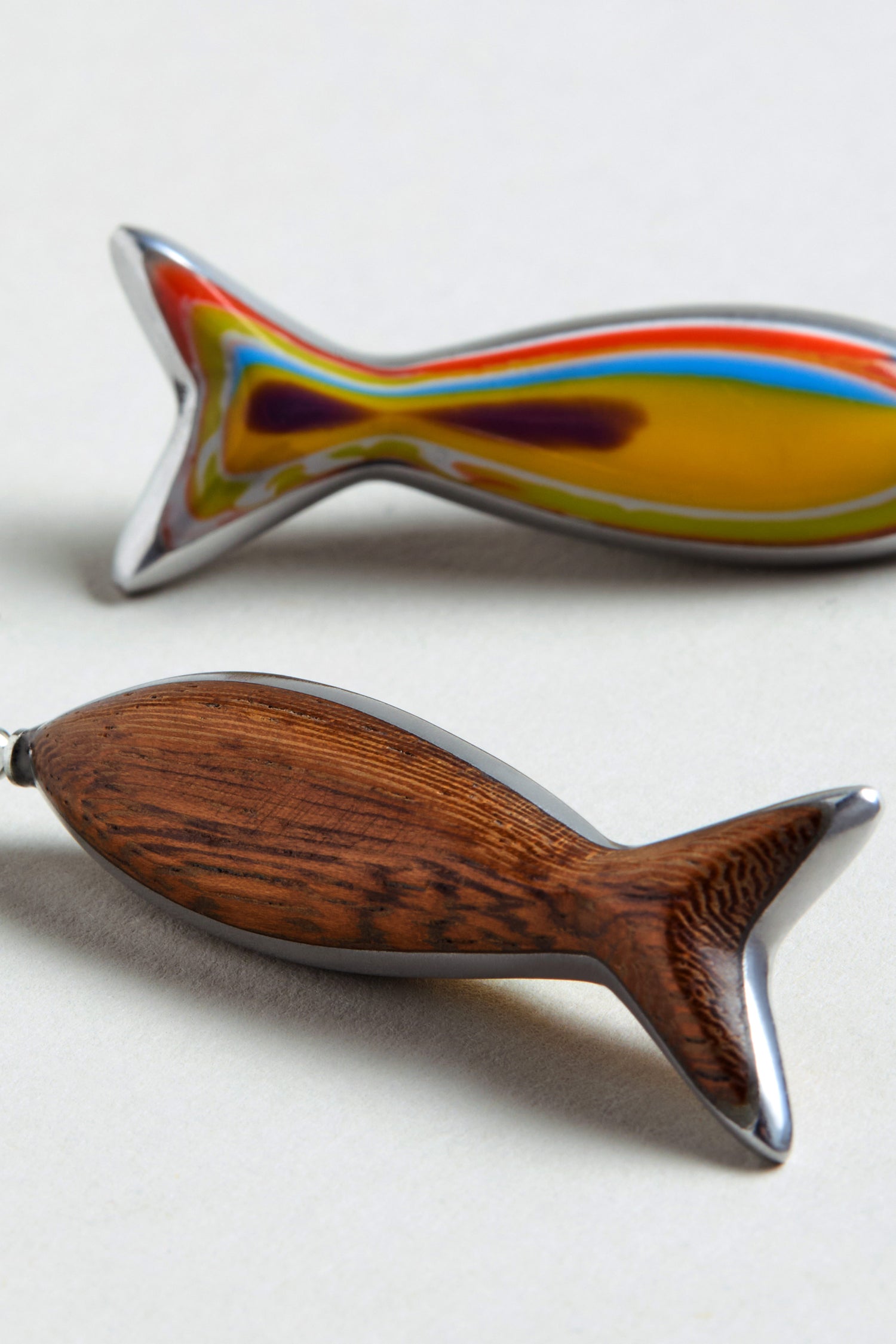 The Poisson Earrings are two fish-shaped recycled aluminium earrings: one with a glossy multicolour resin finish, the other with a wood texture. Both rest on a light background.