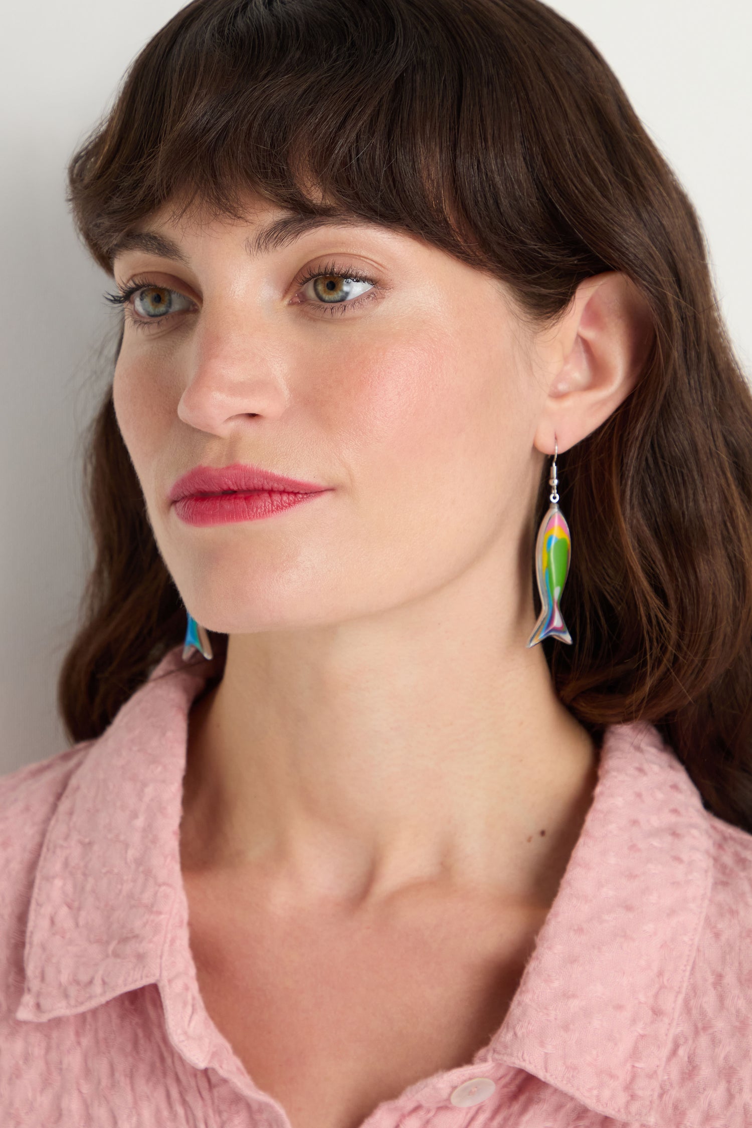 Fair-skinned woman with brown hair wears a pink textured top and vibrant Poisson Earrings—multicolour resin fish shapes—while looking slightly to the side.
