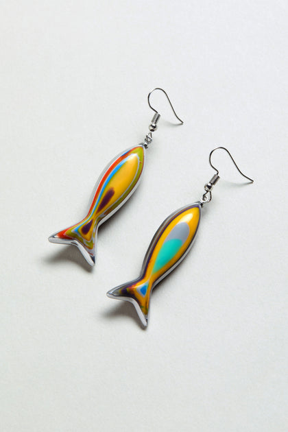 The Poisson Earrings are fish-shaped and crafted from multicolour resin with a vibrant marbled pattern in yellow, orange, blue, and green, shown against a white background.