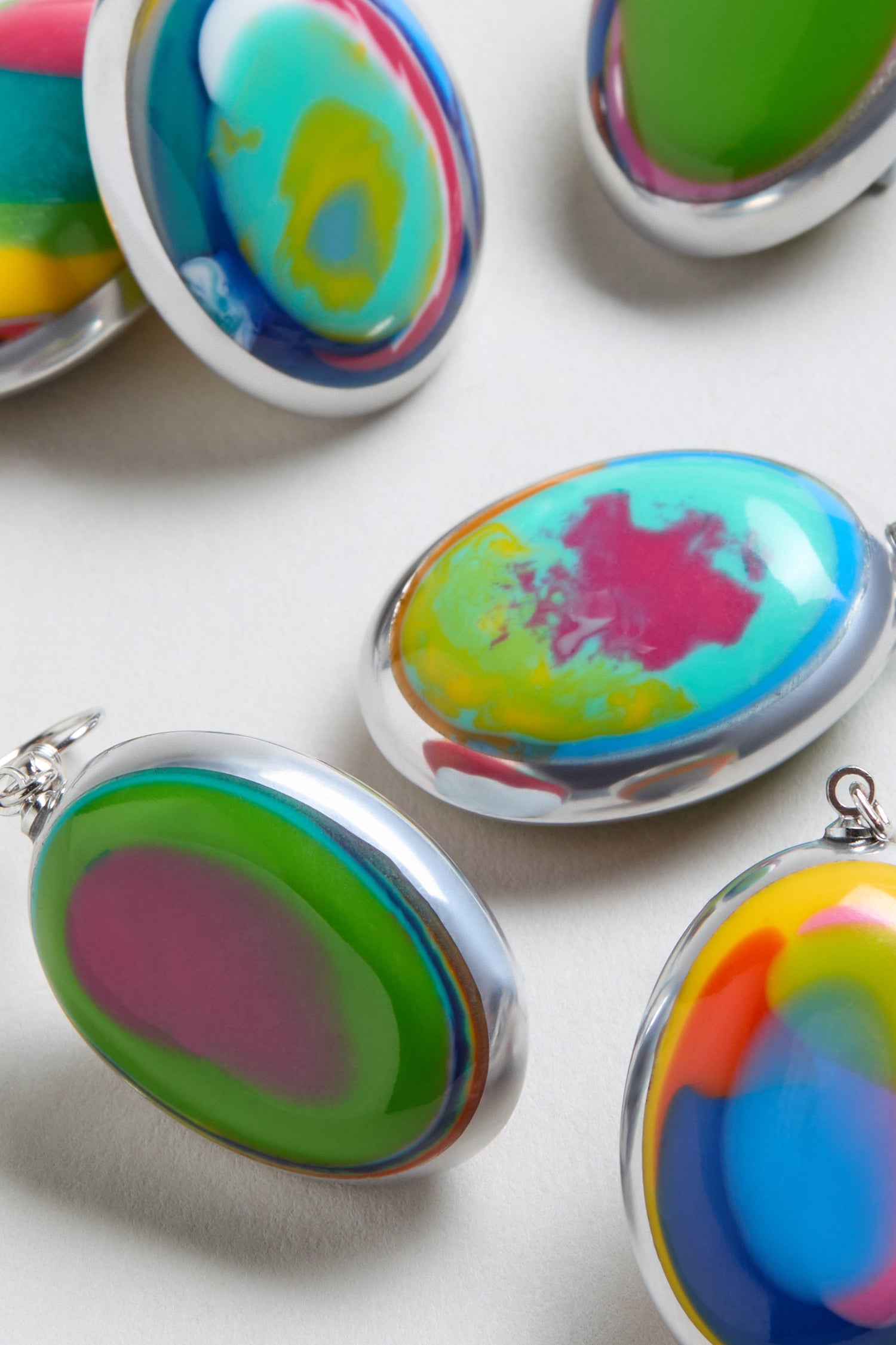 Rainbow Pebble Earrings are oval-shaped with silver edges, featuring vibrant abstract patterns. Arranged on a light surface, their multicolour design is reminiscent of resin jewelry.