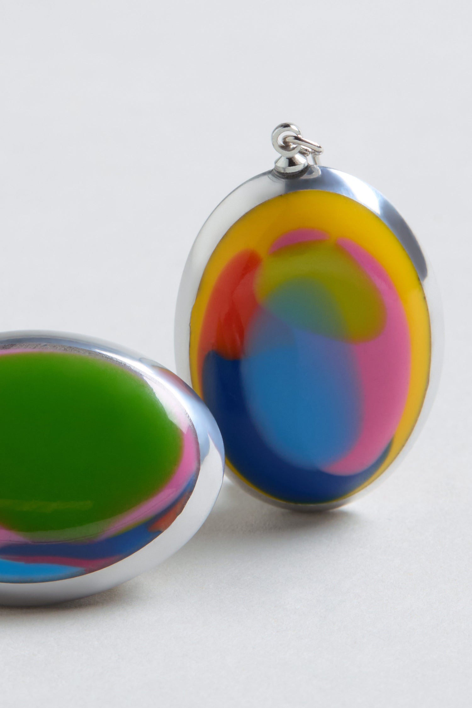Two oval-shaped Rainbow Pebble Earrings, featuring glossy swirled patterns in green, yellow, blue, and pink, are displayed on a light gray surface.