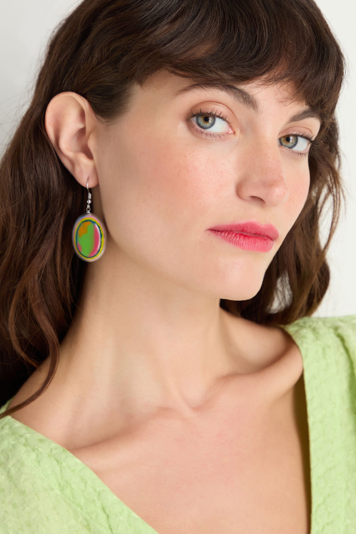 A fair-skinned woman with brown hair wears a green top and Rainbow Pebble Earrings, glancing to the side against a plain background.