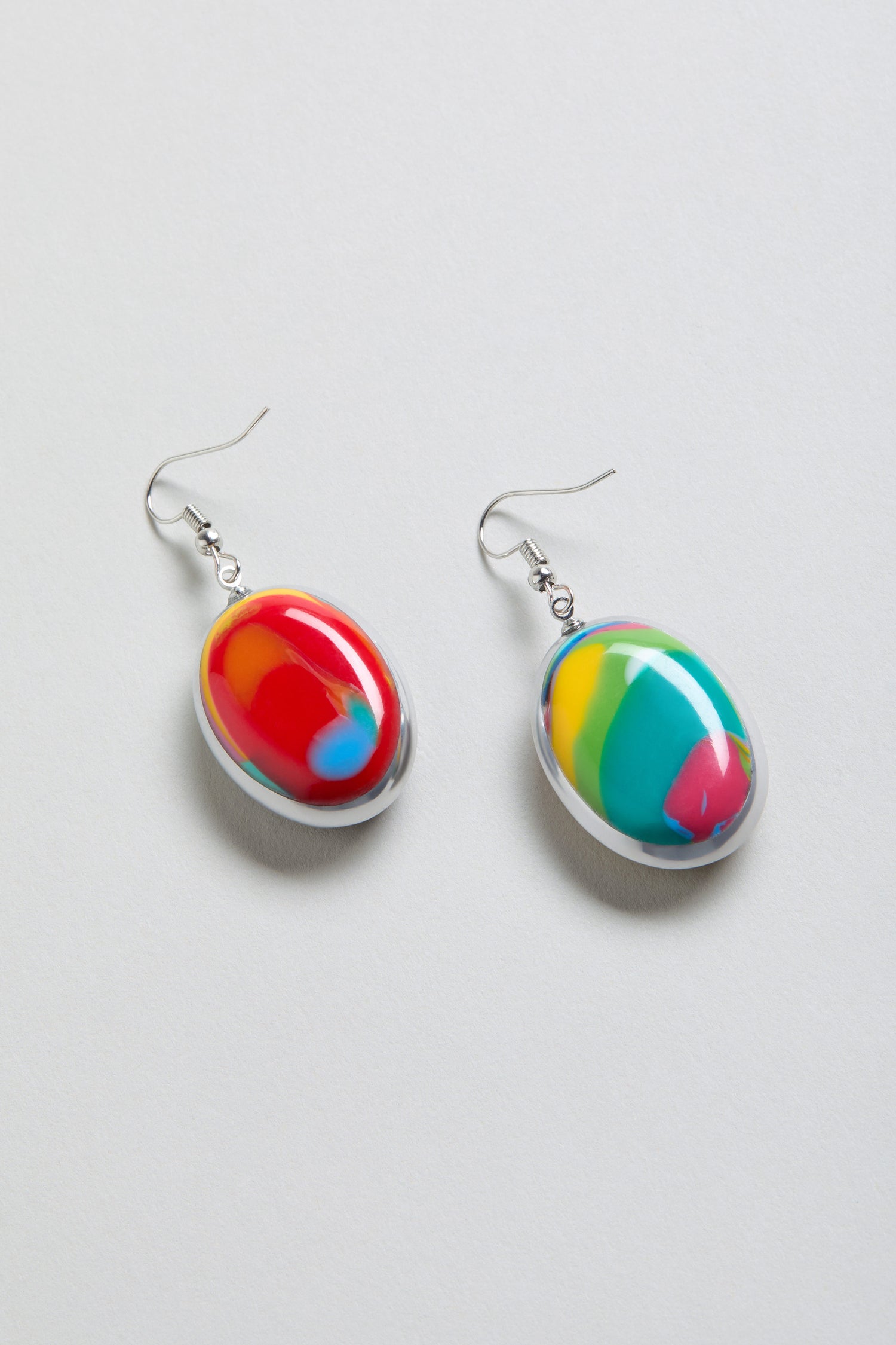 Rainbow Pebble Earrings are oval-shaped, made from multicolour resin with abstract red, yellow, green, and blue patterns, shown on a light background.