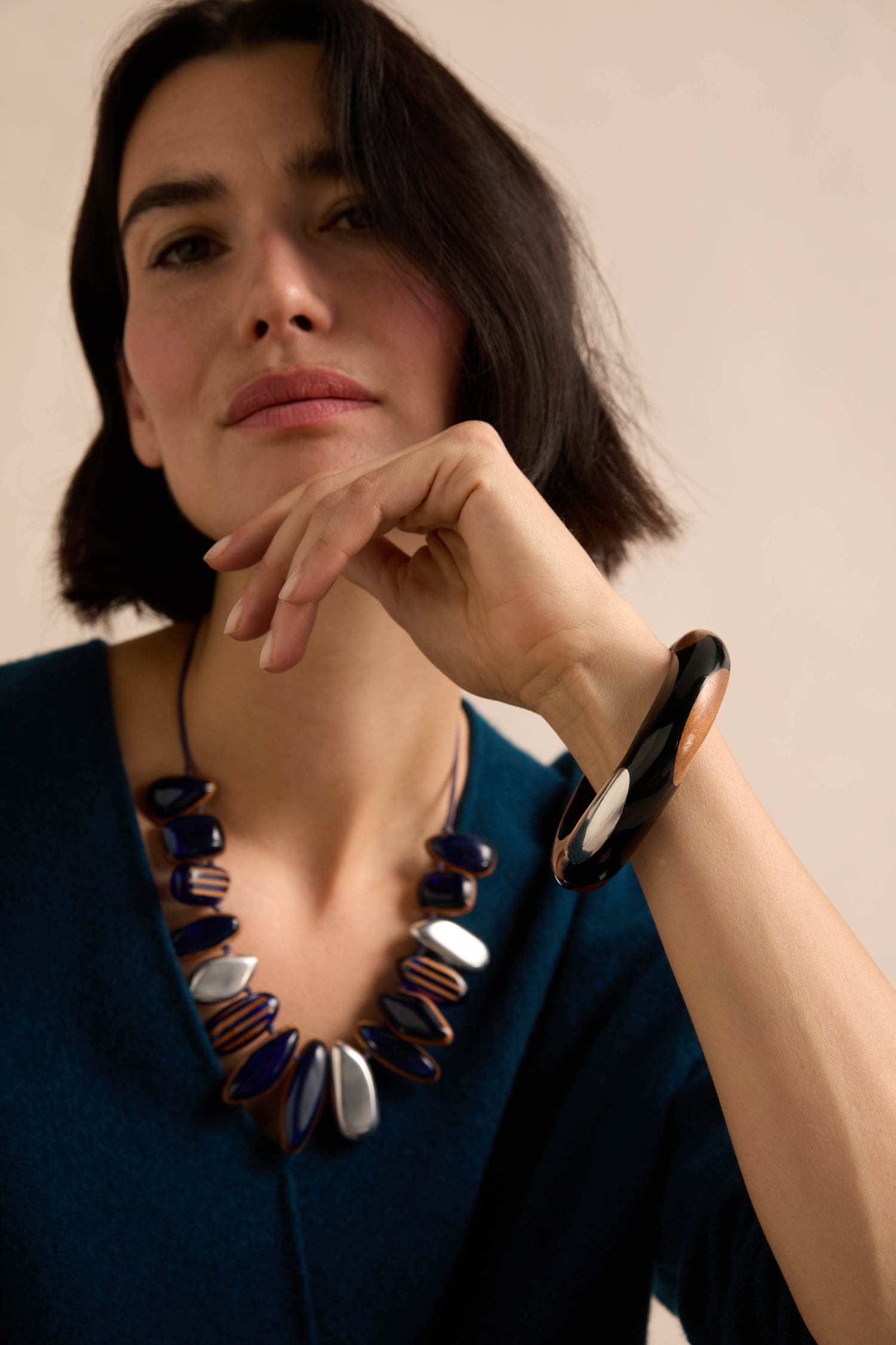 A woman with short dark hair wears a blue top, a chunky dark and metallic necklace, and the striking Dotty Bangle in black wood, resting her chin on her hand as she looks at the camera.