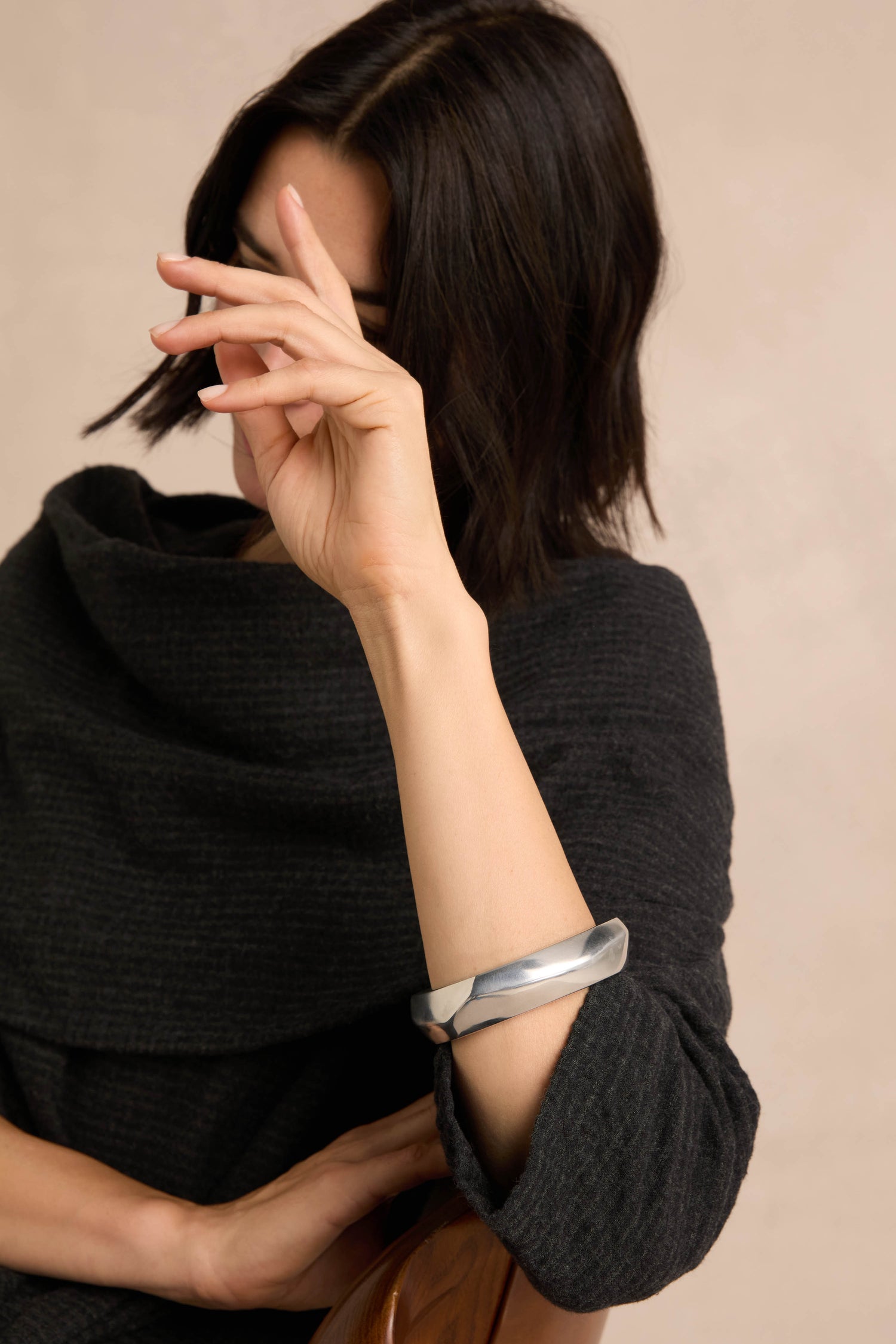 A person with chin-length dark hair wears a black textured top and an architectural jewelry piece—the Statement Silver Bangle—holding their hand up, partially covering their face.