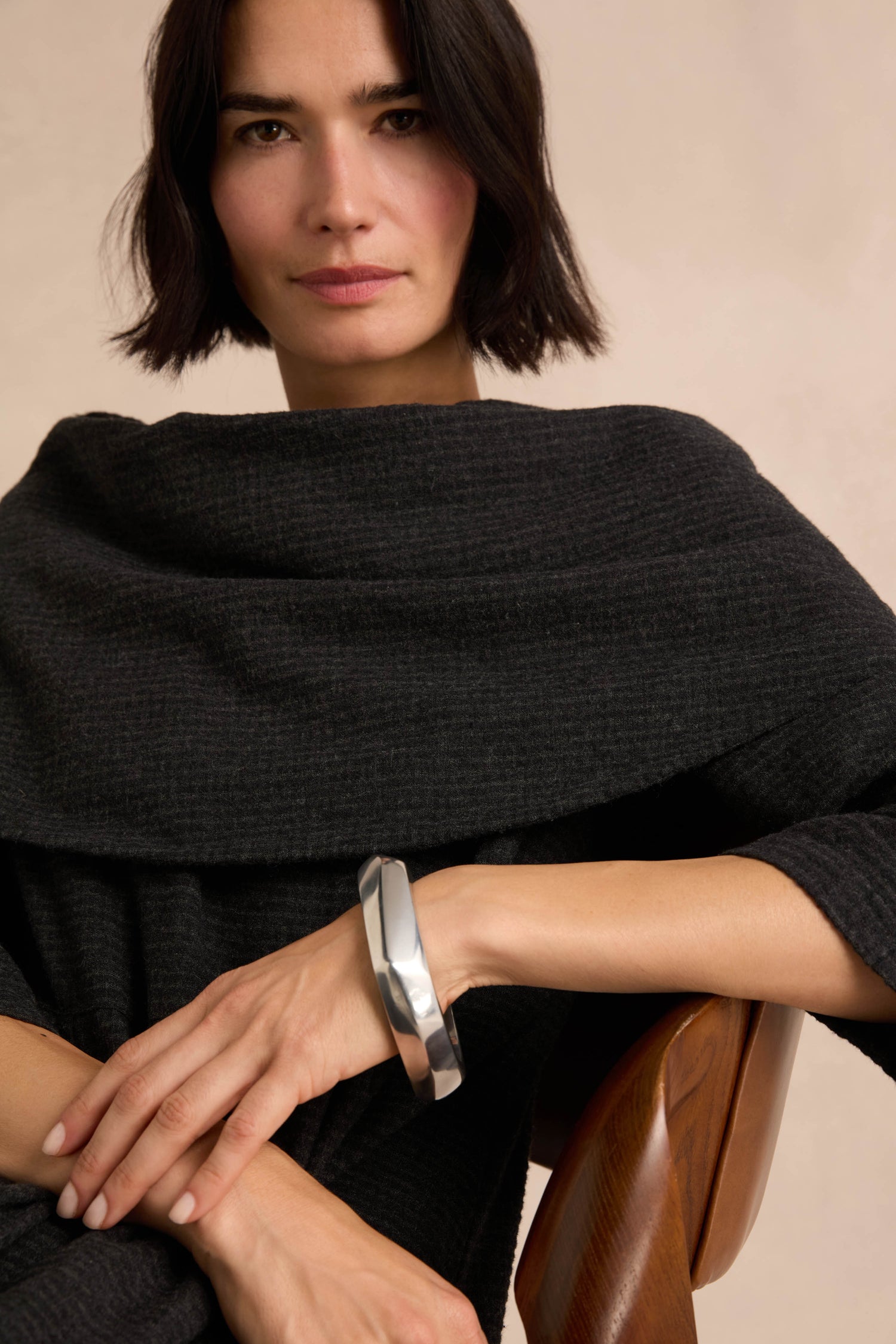 A woman with short dark hair wears a textured dark shawl and the Statement Silver Bangle, a large sculpted silver bracelet, seated on a wooden chair against a neutral background. The architectural design of the Statement Silver Bangle serves as a striking statement piece, highlighting her elegant style.