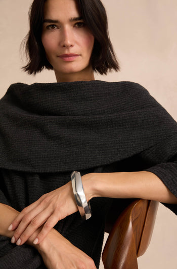 A woman with short dark hair wears a textured dark shawl and the Statement Silver Bangle, a large sculpted silver bracelet, seated on a wooden chair against a neutral background. The architectural design of the Statement Silver Bangle serves as a striking statement piece, highlighting her elegant style.