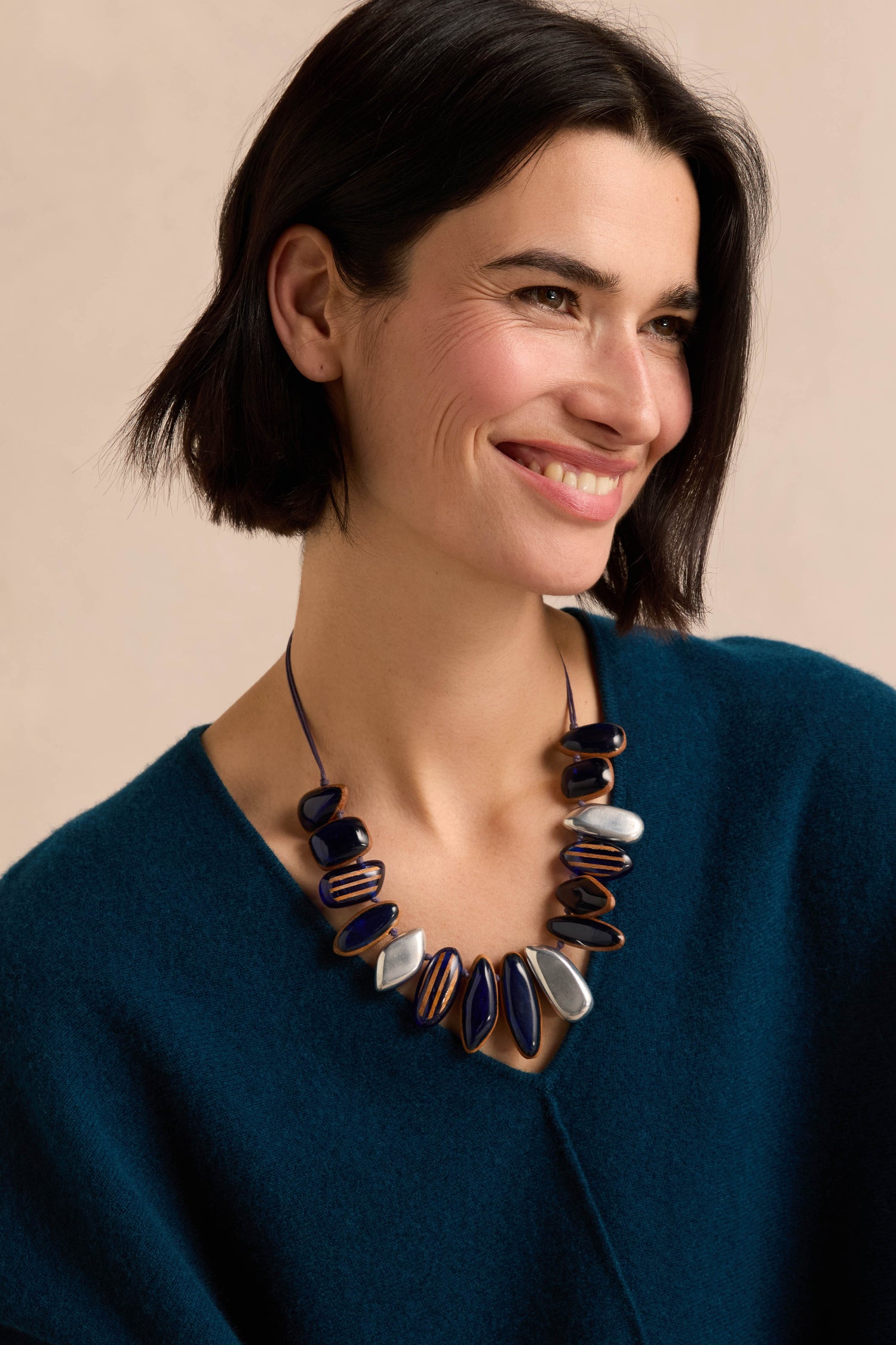 A woman with short dark hair, wearing a teal top and the Resin Pebble Necklace—a chunky accessory featuring blue, brown, and silver resin beads on an adjustable chain—smiles while looking to the side.
