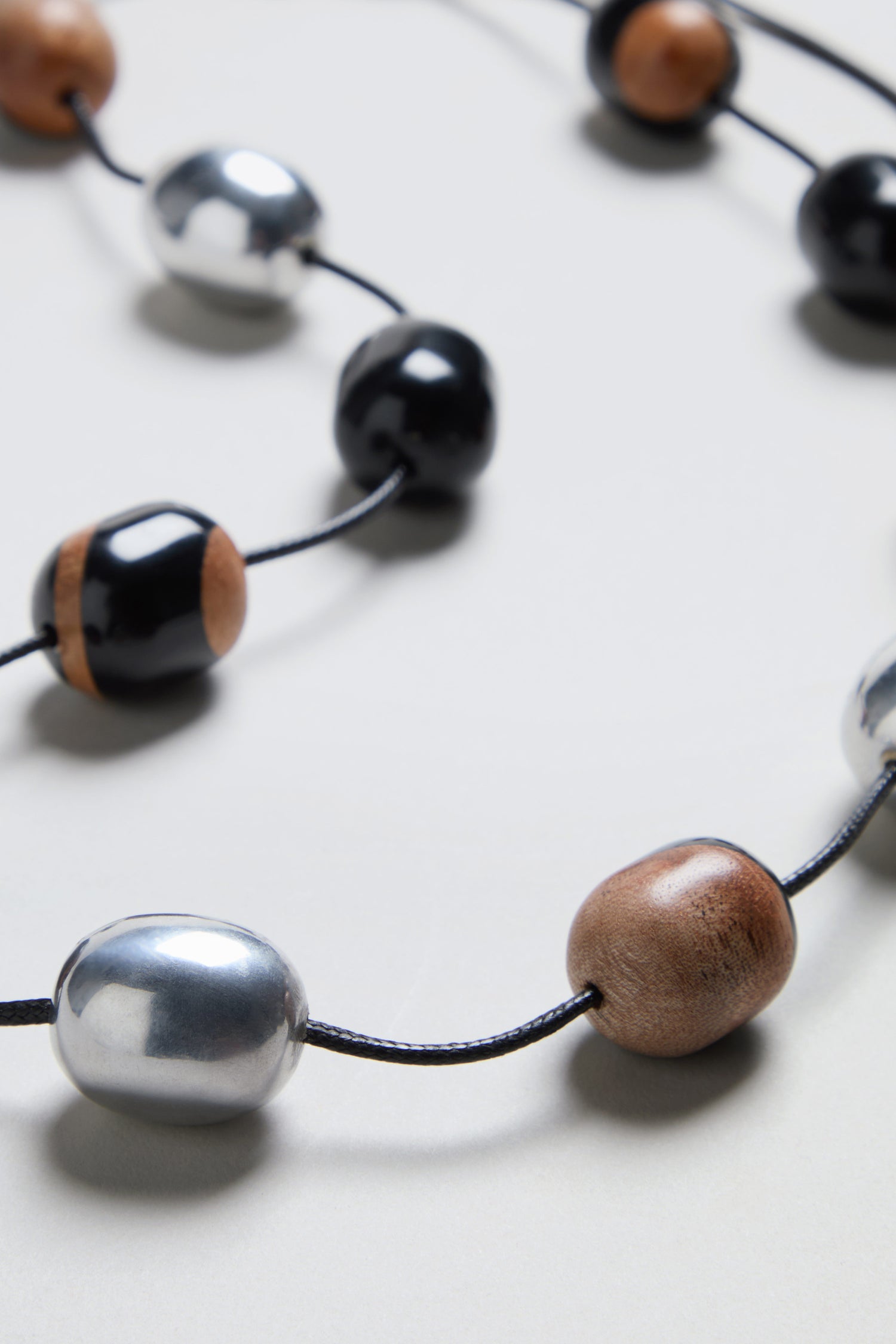 A close-up of the Long Allsorts Necklace, featuring wood and silver elements accented with black, silver, and wood-toned beads elegantly spaced along a black cord resting on a light surface.
