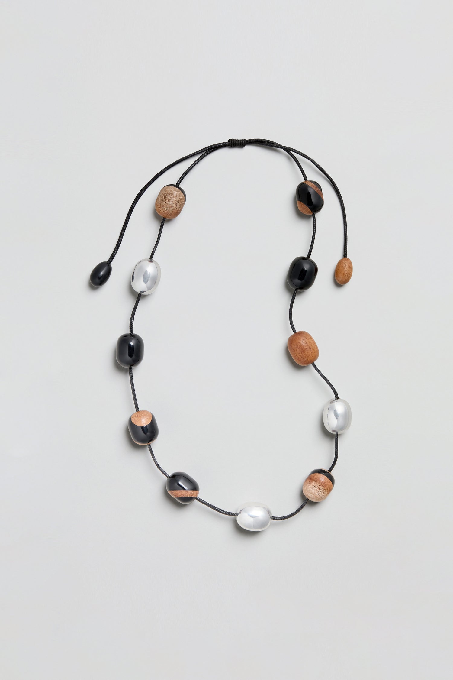 The Long Allsorts Necklace, featuring a black cord strung with alternating black, white, and brown oval beads in a spaced design, is displayed on a light gray background.