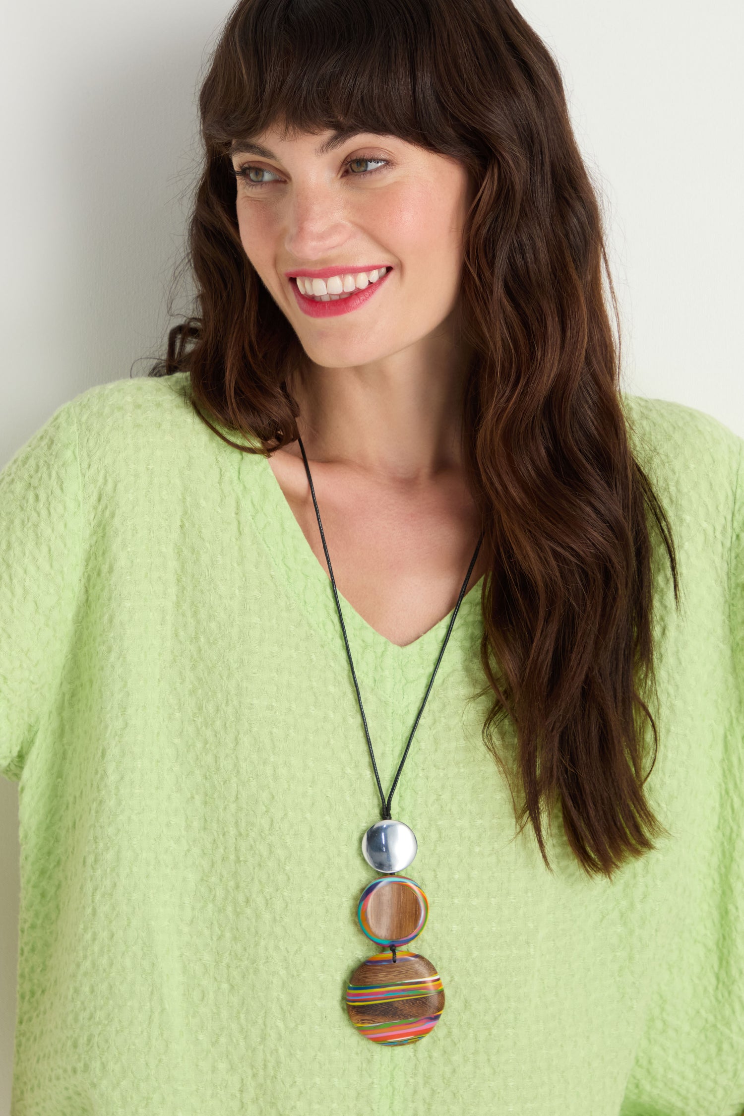 Smiling woman with long brown hair wears a light green textured top and the Colourburst Triple Sphere Necklace, featuring three large circular charms crafted from recycled aluminium, standing against a light background.