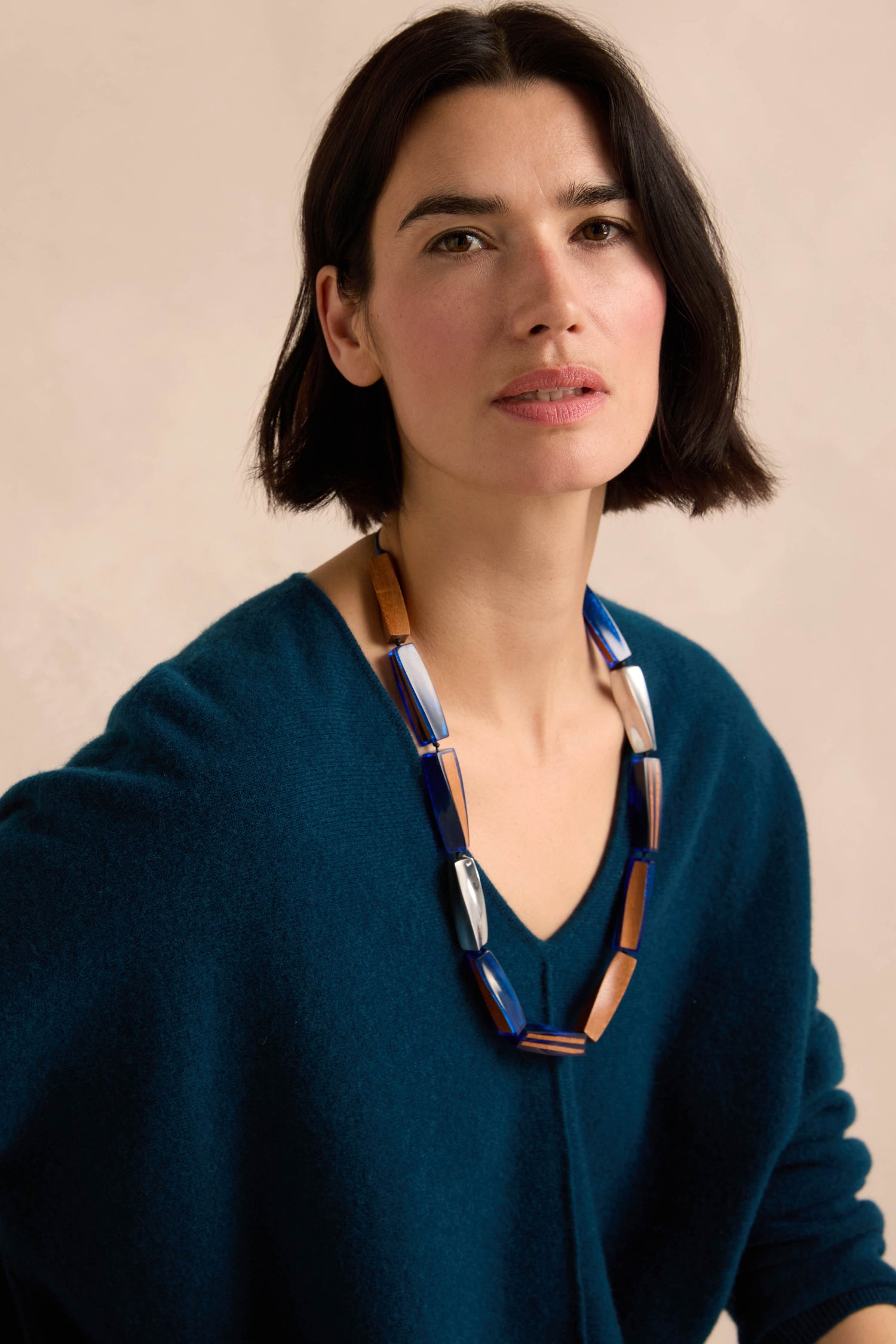 Woman with short dark hair wearing a deep teal sweater and the Twisted Shapes Necklace—an elongated wood accessory featuring chunky geometric elements—looking at the camera against a light beige background.