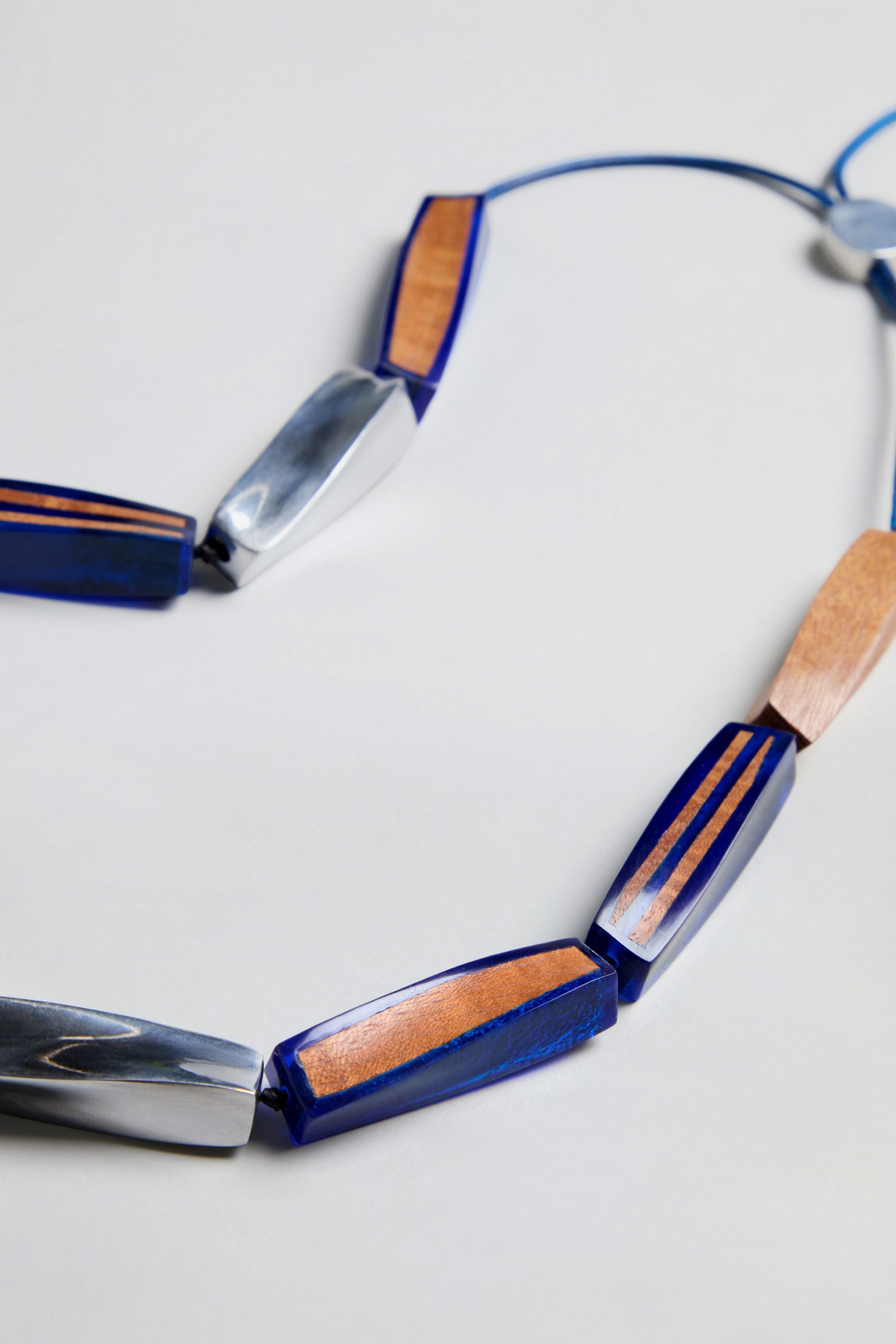 The Twisted Shapes Necklace, showcasing alternating elongated beads in blue, silver, and wood tones, is displayed on a light background.