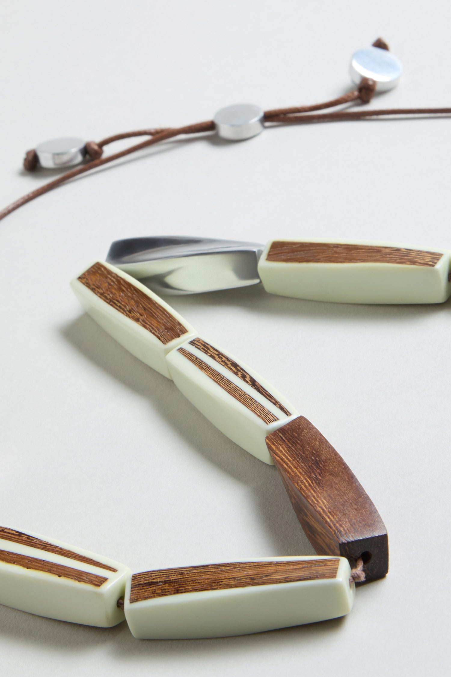 A close-up of the Twisted Shapes Necklace, showcasing rectangular beads with wood and cream inlays on a brown cord, accented by round recycled aluminium.