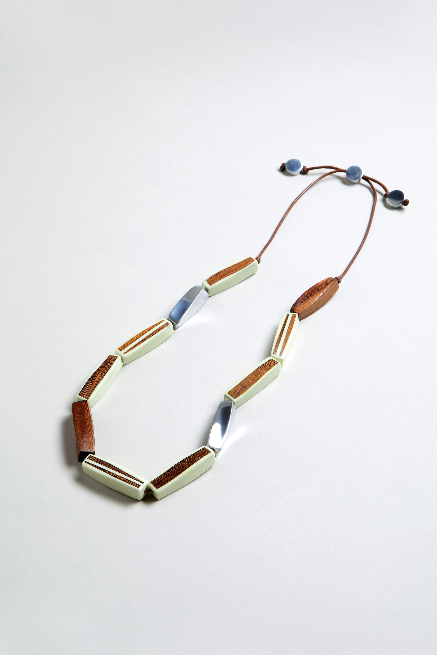 The Twisted Shapes Necklace showcases wooden, white, and recycled aluminum geometric beads on a brown cord with adjustable blue ends, set against a light gray background.