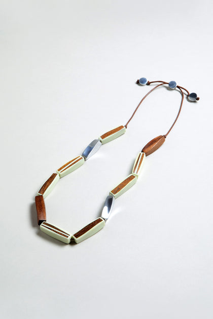 The Twisted Shapes Necklace showcases wooden, white, and recycled aluminum geometric beads on a brown cord with adjustable blue ends, set against a light gray background.