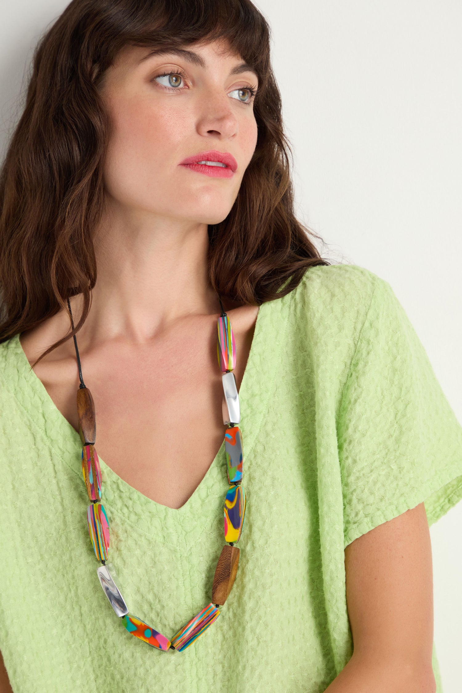 A woman in a textured light green top wears the Twisted Shapes Necklace—featuring colorful beads and recycled aluminium—while looking slightly to the side against a plain background.