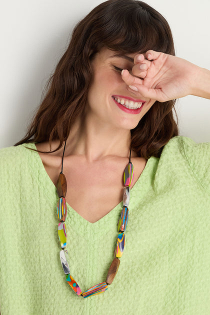 A woman in a light green textured top smiles with her eyes closed, partly hiding her face with her hand, wearing the Twisted Shapes Necklace crafted from recycled aluminium and natural wood beads.