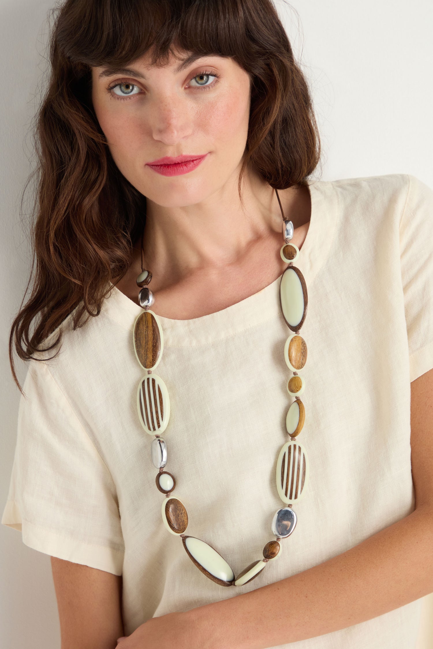 Woman with wavy brown hair wears a cream top and the Humbug Necklace, which features oval and round beads crafted from recycled aluminium and natural wood tones, standing against a plain background.