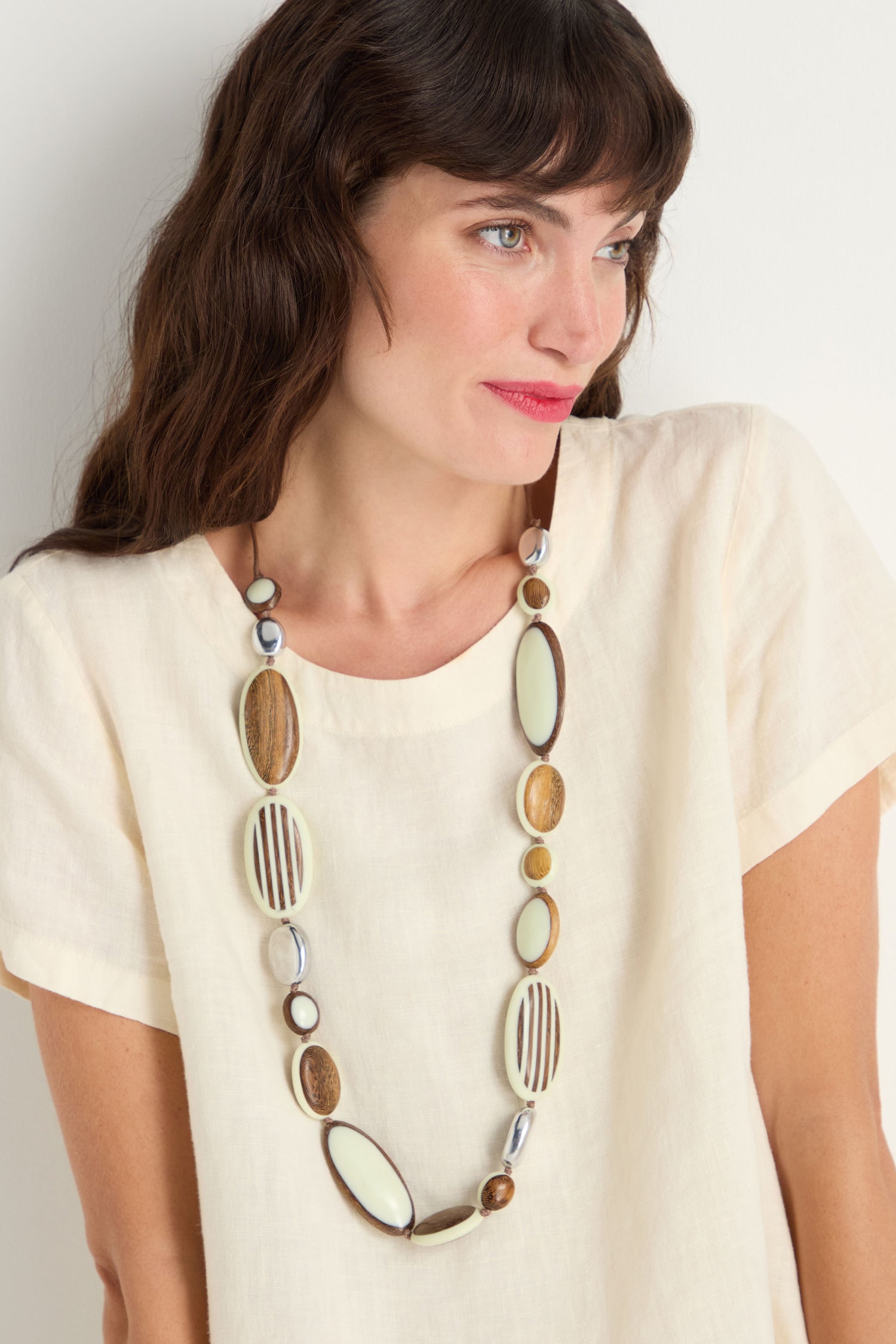 Woman with long brown hair wearing a cream short-sleeve top and the Humbug Necklace—a beige resin accessory with oval and round beads in natural tones.