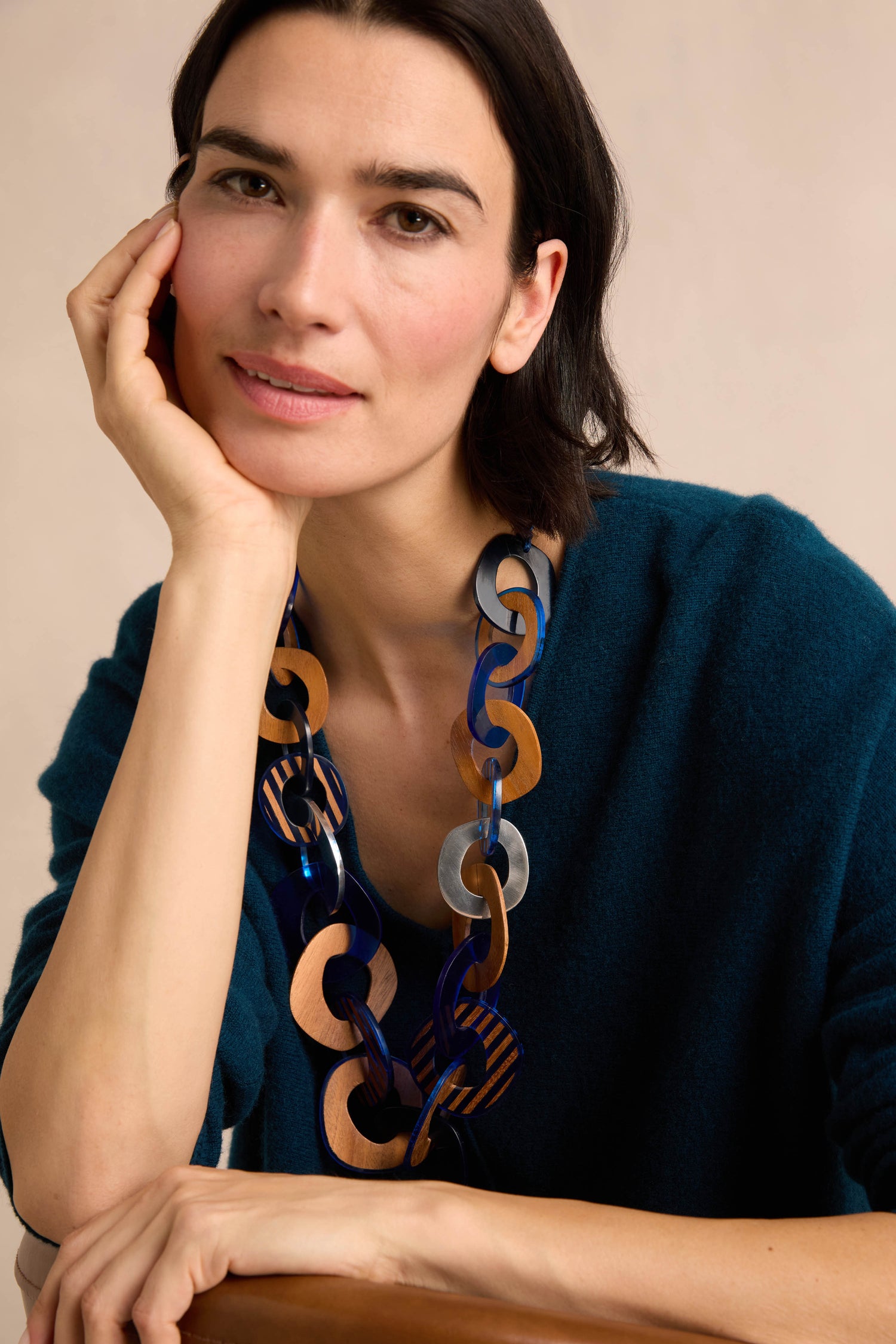 A woman with short dark hair wears a teal sweater and the Blue Stripe Wooden Resin Links Necklace—a striking statement piece featuring large, linked elements in brown, blue, and silver tones with bold resin and wood accents—as she sits with her hand resting on her face.