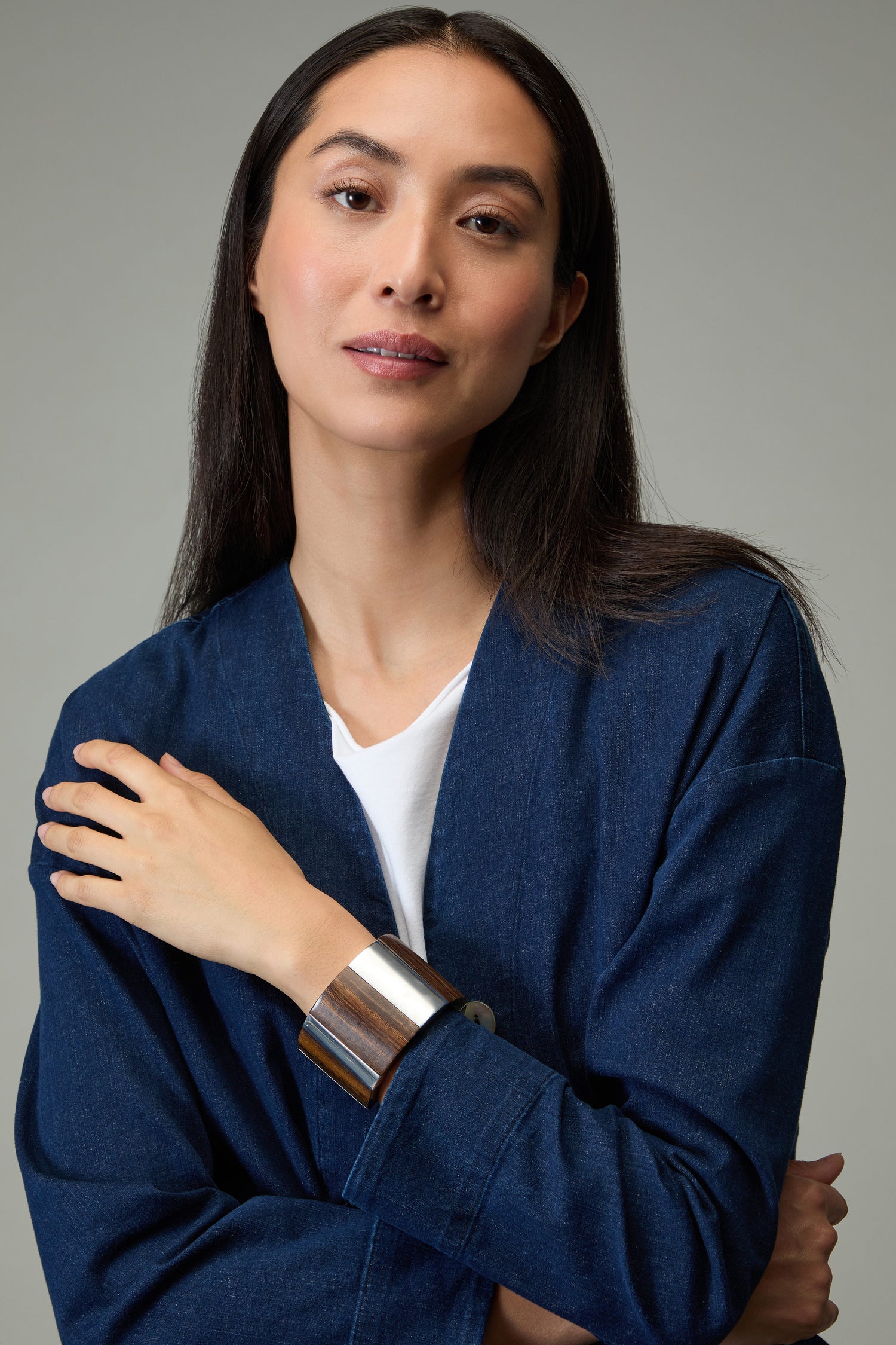 A woman with long dark hair, in a blue denim jacket and white top, poses with one arm across her chest against a neutral background while wearing the Textured Wooden Bracelet.