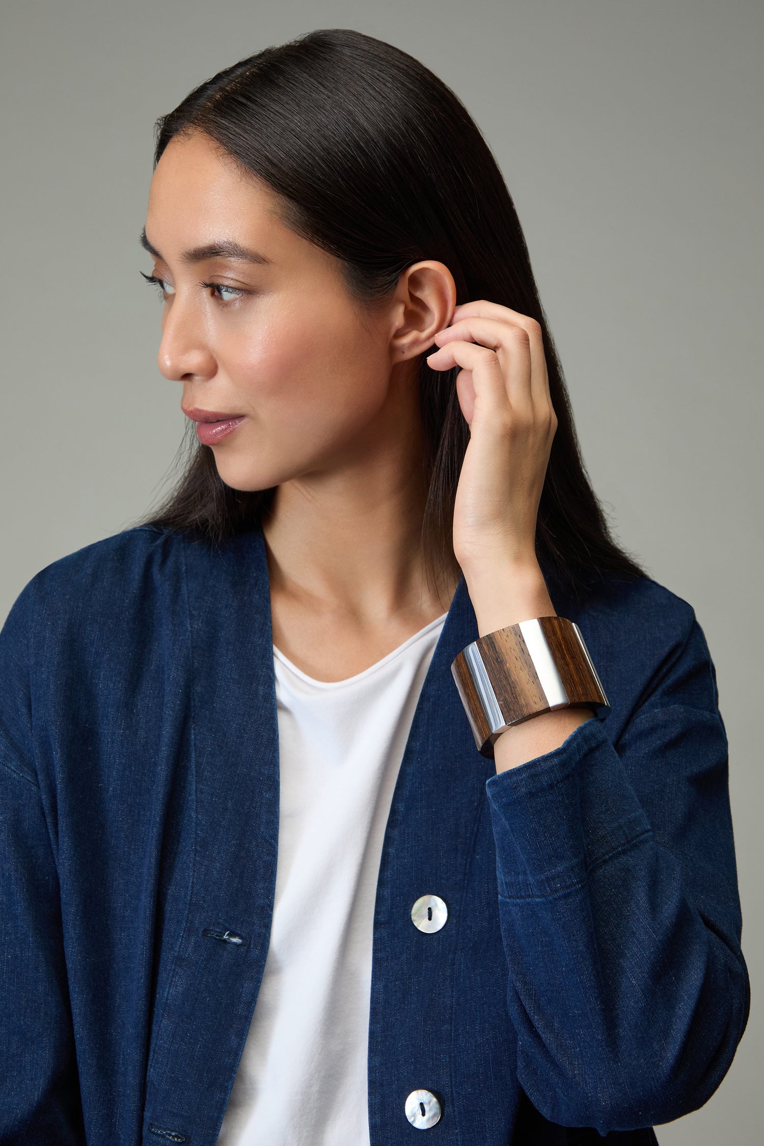 A woman with long dark hair in a blue jacket and white shirt looks to the side, touching her ear. She wears a large Textured Wooden Bracelet on her wrist.