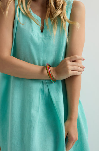 A person in a sleeveless turquoise dress stands with crossed arms, wearing a sculptural bangle and the Flores Rainbow Bangle on one wrist.
