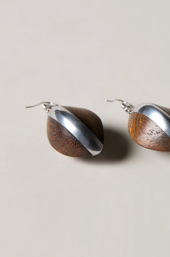 The Wooden Oval Pebble Earrings showcase rounded wooden shapes accented with diagonal silver metal, elegantly presented against a light background.