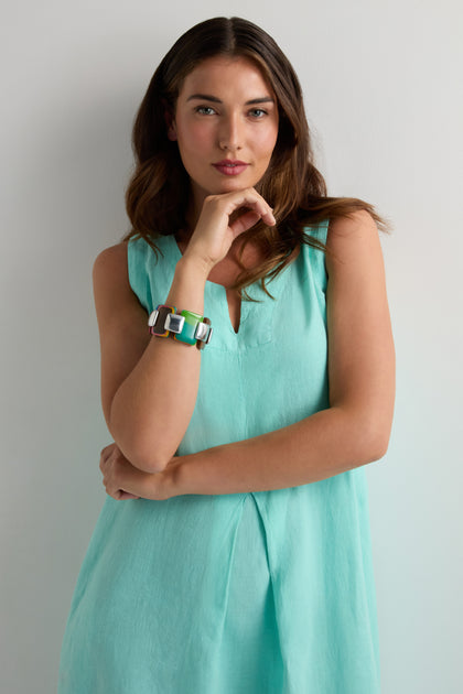 A woman in a sleeveless turquoise dress stands against a plain wall, wearing the Colourburst Squares Links Bracelet—a chunky handcrafted marbled resin piece inspired by Barcelona—and rests her hand on her chin.