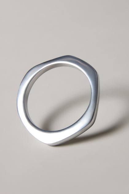 The Solar Silver Bangle is a handmade Spanish bracelet featuring a silver, geometric design with a slightly irregular, rounded polygon shape—an elegant accessory casting a subtle shadow on a neutral background.