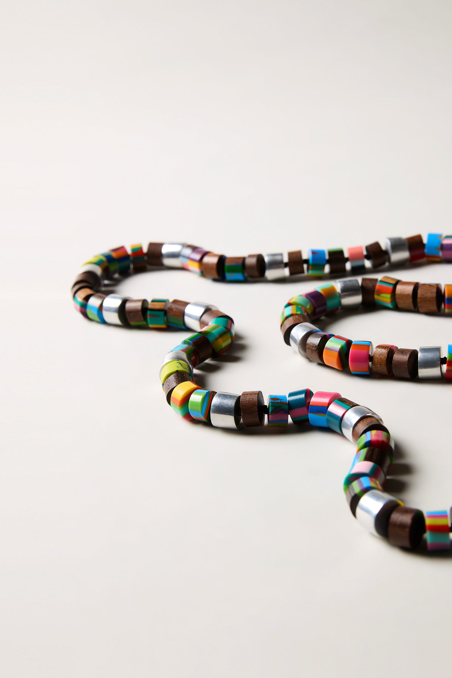 The Venus Colourburst Cylinders Necklace features handcrafted alternating wooden and colorful metallic beads, artfully arranged on a simple surface.