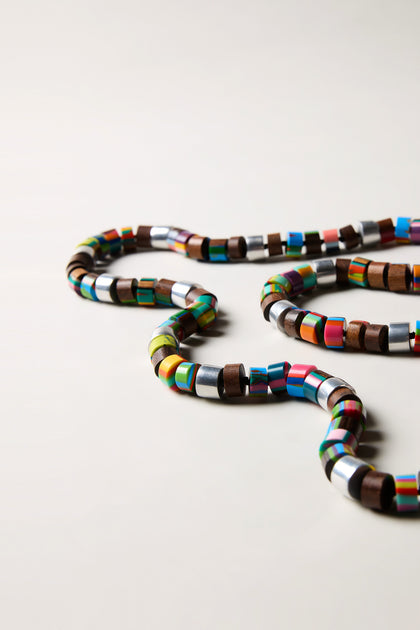 The Venus Colourburst Cylinders Necklace features handcrafted alternating wooden and colorful metallic beads, artfully arranged on a simple surface.