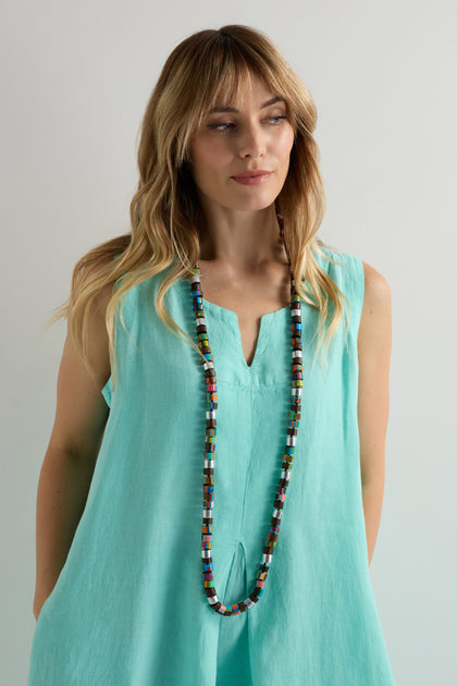 A woman with long blonde hair in a sleeveless turquoise dress wears the Venus Colourburst Cylinders Necklace, standing against a plain light background.