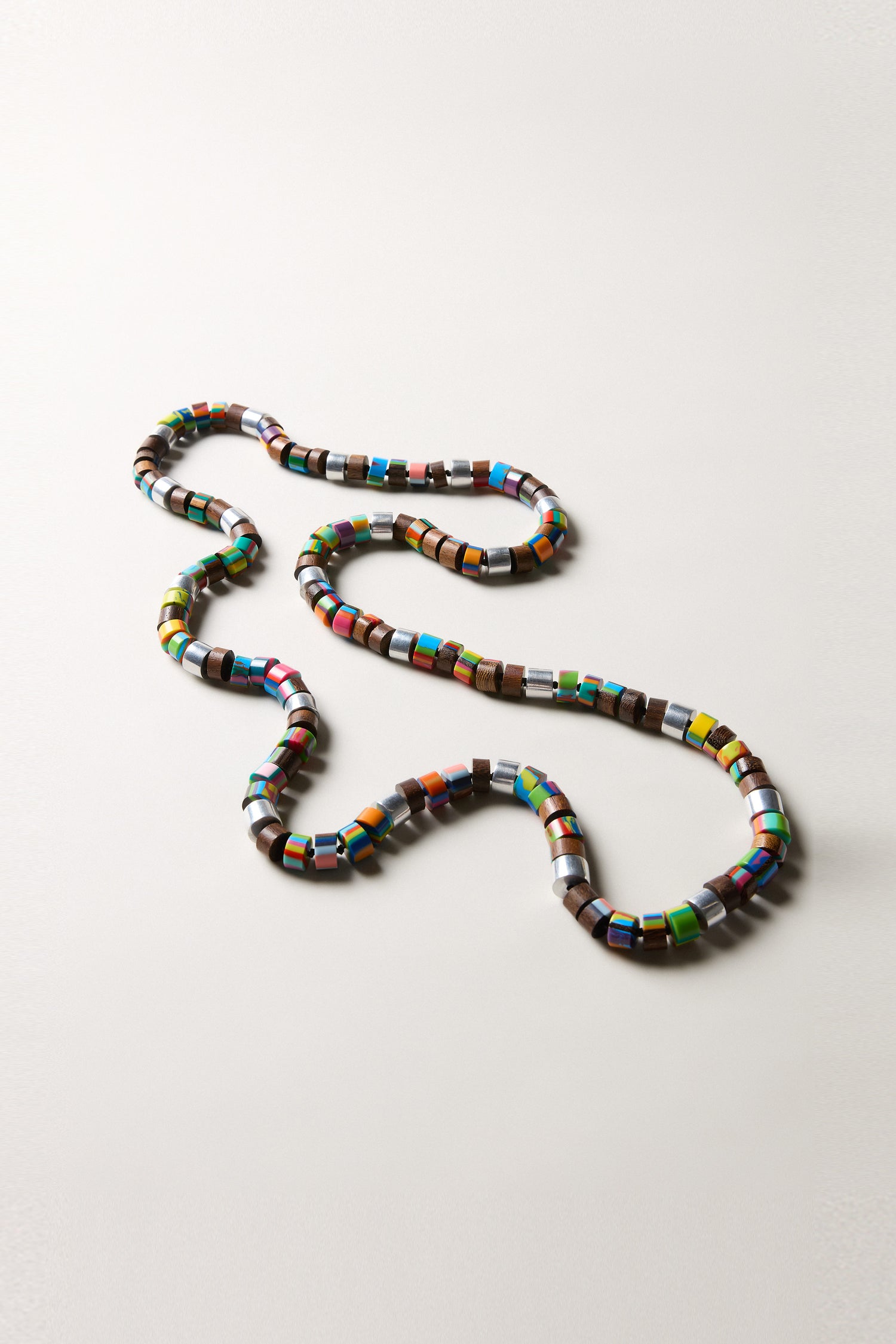 The Venus Colourburst Cylinders Necklace features handcrafted, multi-colored cylindrical beads arranged in a wavy pattern on a plain, light background.