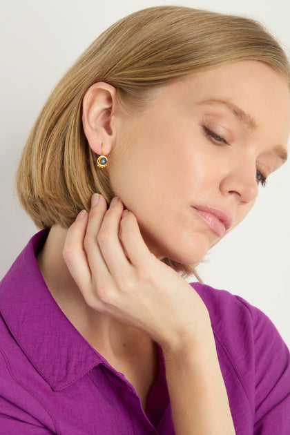 A woman with short blond hair, dressed in a purple collared shirt, touches her neck and looks downward while wearing Labradorite Stone Earrings.