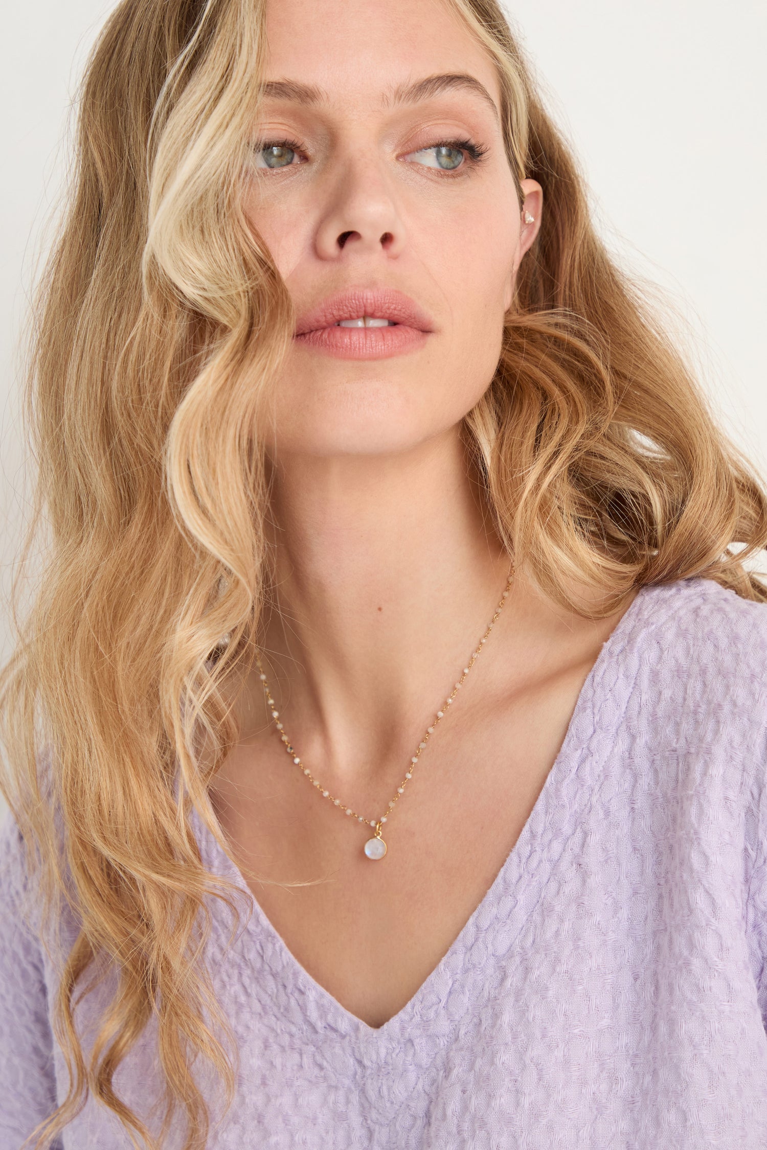 Woman with long blonde hair wearing a textured light purple top and a delicate Hand Crafted Moonstone Bead Necklace, featuring a round pendant, looking slightly to the side.