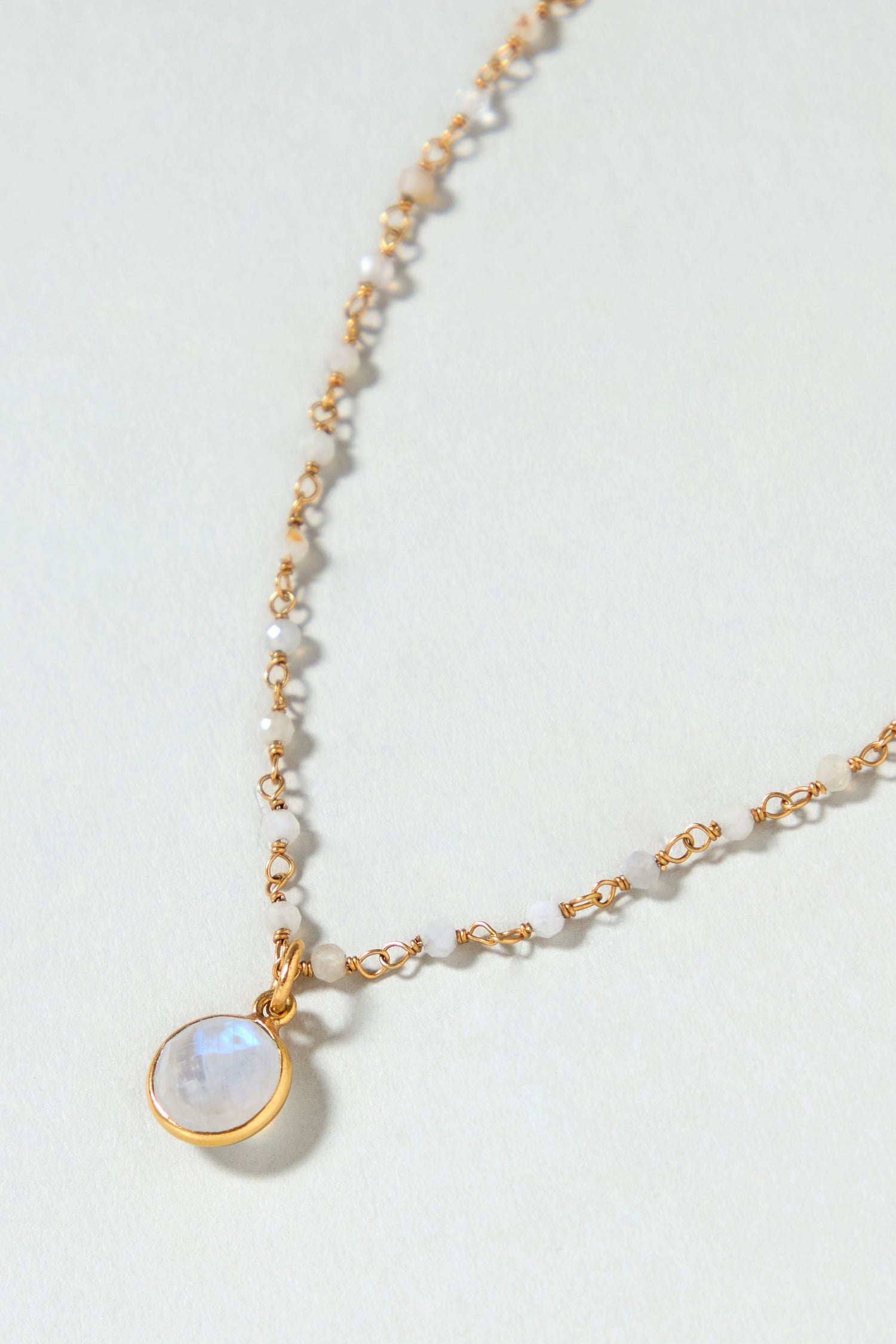 The Hand Crafted Moonstone Bead Necklace features gold plating, small white beads, and a round iridescent moonstone pendant, shown against a light background.
