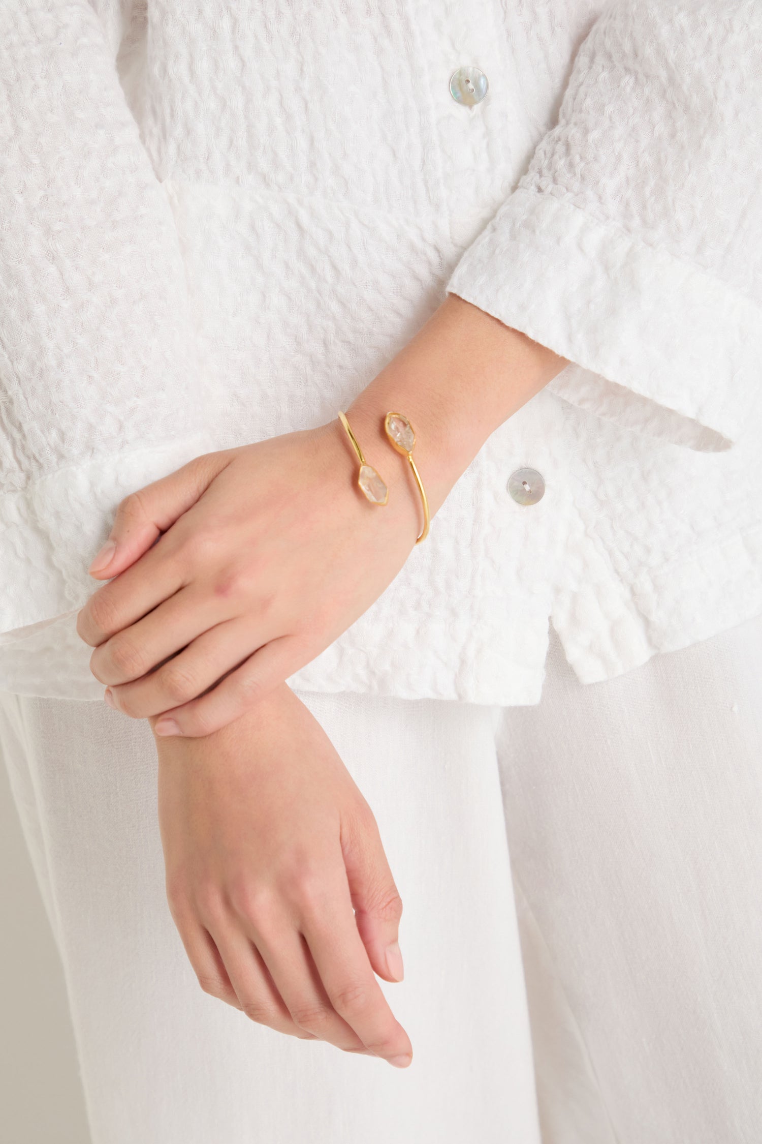 A person wears a white textured outfit with the Gold Plated Quartz Herkimer Diamond Bracelet—an 18k gold plated open cuff adorned with two sparkling quartz Herkimer diamond accents—on their wrist.